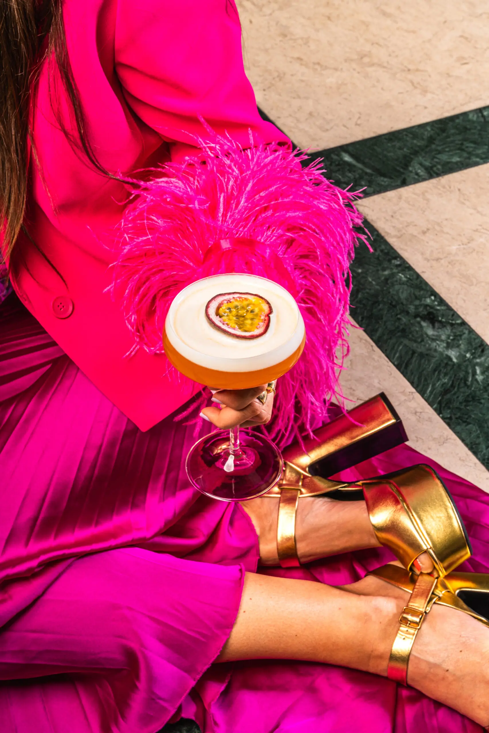 Een dame in felroze kleding met feestelijke gouden hakken die een Pornstar Martini vasthoudt
