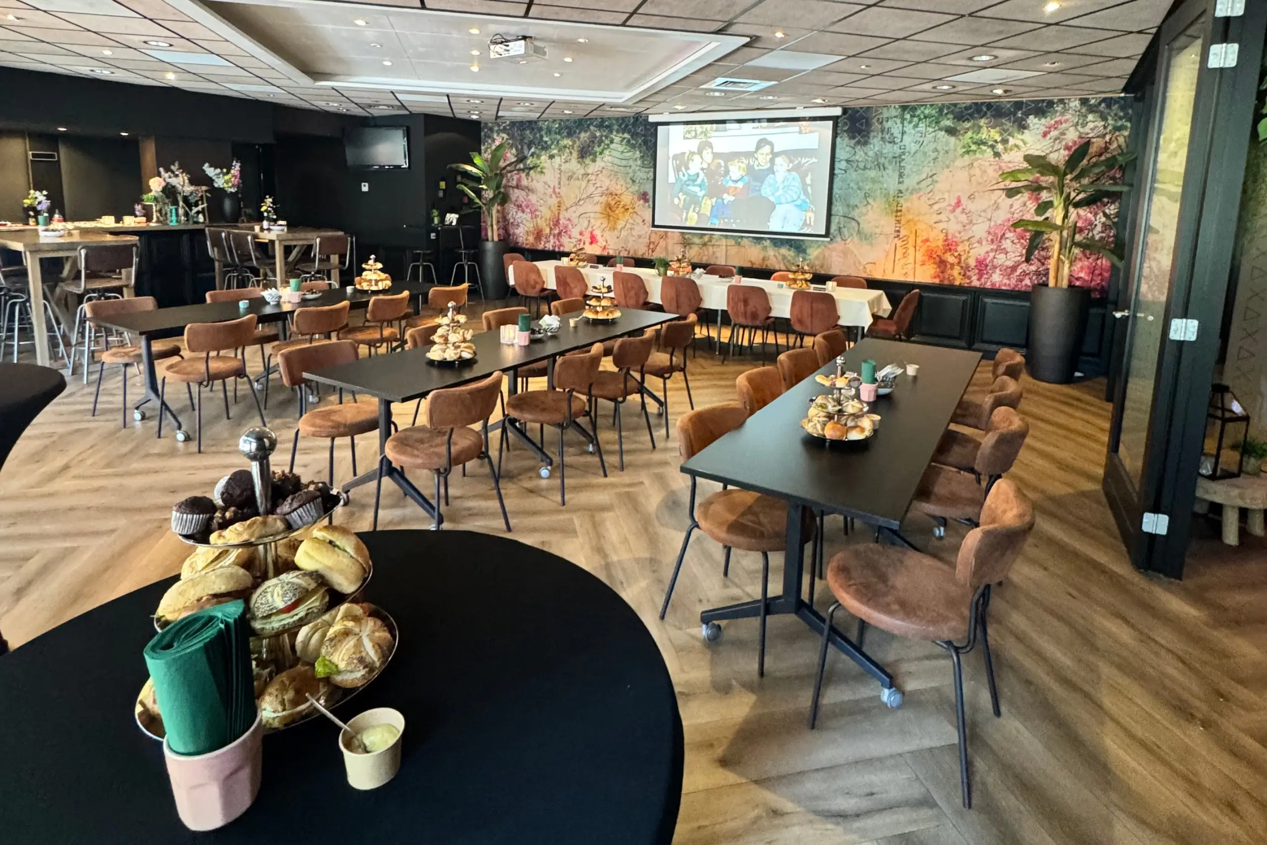 Ruime feest- en vergaderzaal met gedekte tafels, bruine stoelen, hapjes en een groot projectiescherm.