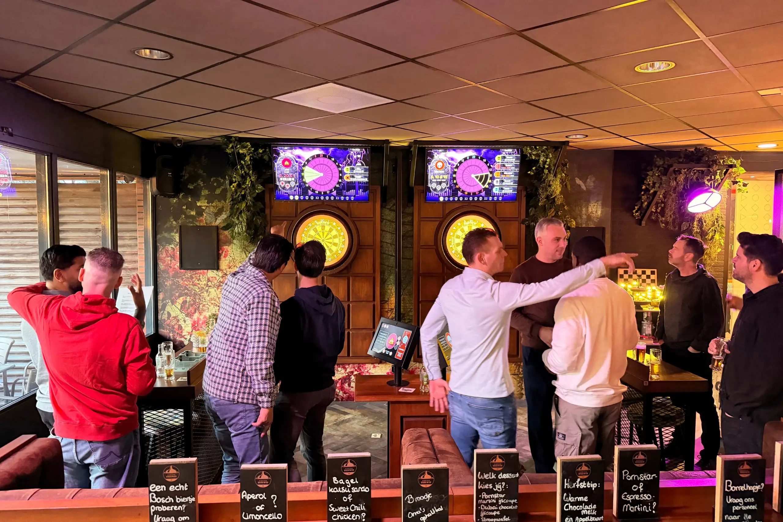 Groep mannen speelt interactief darts op twee digitale dartbanen in een kleurrijk verlichte ruimte.